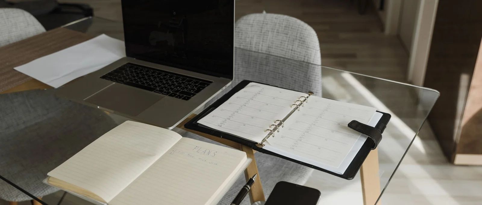 Laptop, open notebook and planner arranged on a glass desk